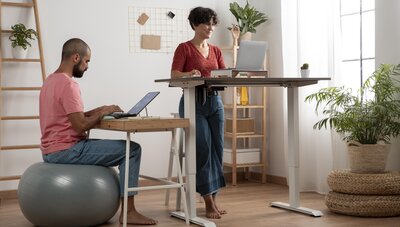 Frau und Mann im Homeoffice. Frau steht an einer Stehlösung. Mann sitzt auf einem Sitzball am Schreibtisch.
