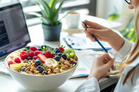 Frau sitzt am Schreibtisch und hat eine große Schale mit Müsli und Obst vor sich